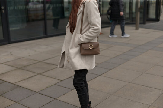A woman dressed in a stylish white coat paired with black pants is standing on a bustling sidewalk, surrounded by pedestrians and various shops