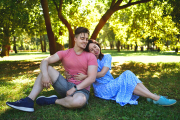 A mother and her adult son are having a rest in nature, sitting on the green grass against the backdrop of trees.