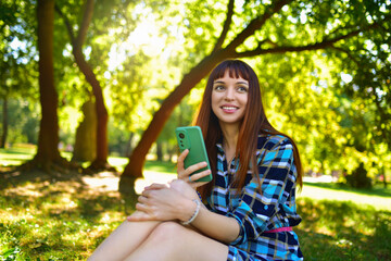 A young woman sitting in nature on the green grass, looking at her phone against the background of the spring park.