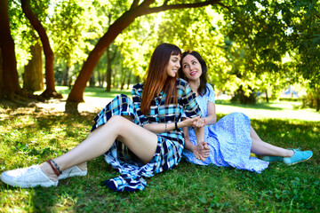 Two ladies resting on the grass in nature and talking to each other with smiles against the background of the spring park.