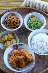 Indonesian cuisine served on a bamboo tray. rice, stir-fried chayote, stir-fried tempeh, balado chicken, fried tofu, crackers. Tumis labu siam, ore tempe, Tahu goreng, ayam bumbu pedas.
