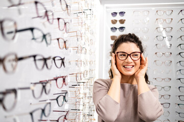 Beautiful brunette girl trying on new eyeglasses frame in optician shop.