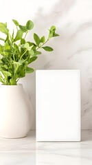 A blank white canvas stands upright on a white marble surface, with a vase of green leaves in the foreground