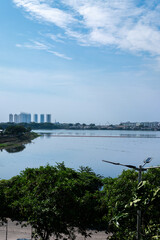 Fototapeta premium A calm lake reflects the sky and distant buildings, framed by green foliage. Serene and expansive.