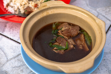 A clay pot of rich, dark Bak Kut Teh (pork rib soup) with tender pork ribs and leafy greens, served on a blue saucer. Flavorful Southeast Asian herbal broth.