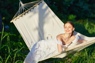 Young woman in a white dress relaxing in a hammock in a green garden, smiling joyfully, with soft...