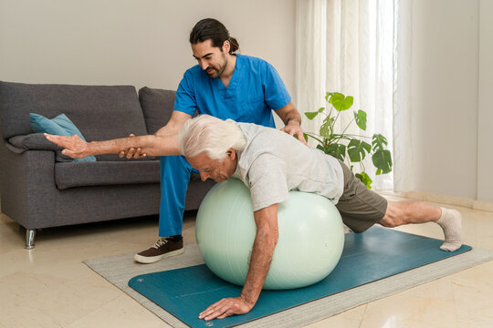 Senior man doing physiotherapy exercises with fitness ball and personal trainer at home