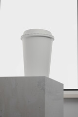mockup of paper coffee cup standing on cement podium in front of glass window.