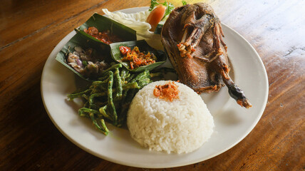 Delicious Balinese crispy fried duck from Bebek Tepi Sawah Restaurant, served golden brown with rice, various vegetables, and spicy sambal on a white plate with wooden background. Close up