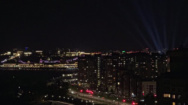 Urban nightscape with glowing city lights, traffic movement and distant searchlight beams illuminating the skyline over modern high-rise buildings and historic architecture