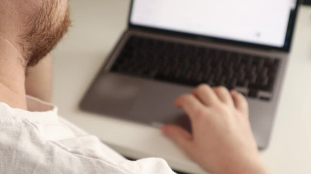 A man uses the trackpad on his laptop to surf the internet sitting at a white minimalist desk