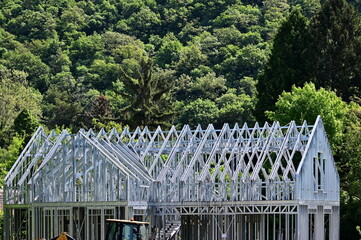galvanized metal stud exterior wall frame for residential building. sloped roof. openings for windows. house construction process. lush green background. trees on a hill side. modern frame structure