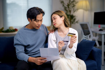 Young asian couple calculating domestic bills and having financial problems holding credit cards sitting on sofa at home