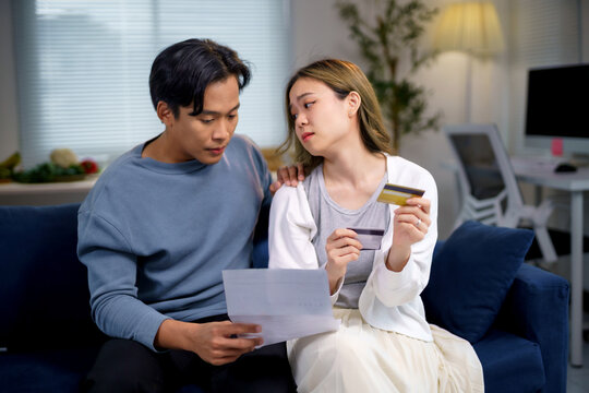 Young asian couple calculating domestic bills and having financial problems holding credit cards sitting on sofa at home - Powered by Adobe