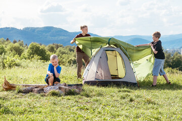 Three brothers have picnic. Boy roasting sausages on sticks over campfire flame while two brothers set up the green tent. Happy family outdoor picnic camping activities concept