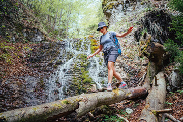Active woman with backpack in hat dressed in active trekking clothes crossing mountain river creek with waterfall by fallen tree trunk bridge. Traveling, trekking, nature concept image