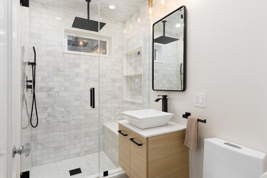 A bathroom with a floating wood cabinet, vessel sink on white marble countertop, black faucets, and a shower with marble subway tiles.