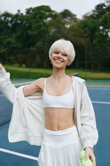 Fotobehang Cheerful young woman with short blonde hair in sporty outfit, smiling on tennis court with green background, embodying a healthy lifestyle and vibrant energy © SHOTPRIME STUDIO