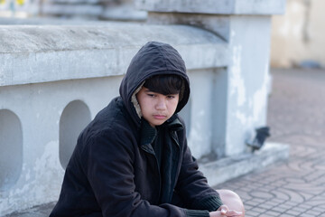 Asian young boy wearing a dark hooded jacket, sitting near a stone fence or barrier thoughtfully...