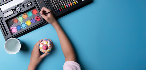 Kid hands painting color on the plaster cake statue at the table with art supplies, top view