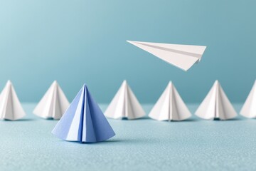 Naklejka premium Outliers Differences concept. A single blue origami paper among white ones, with a paper plane flying above, symbolizing leadership and individuality.