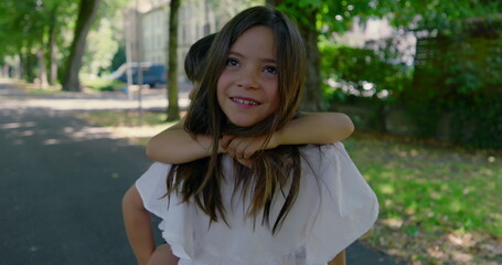 Sister giving her younger brother a piggyback ride on a sunny day, both laughing and enjoying the outdoor park, surrounded by greenery and a shaded walking path