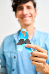 Cheerful Latin man showing blue ribbon with mustache. Prostate cancer, awareness concept, men Health