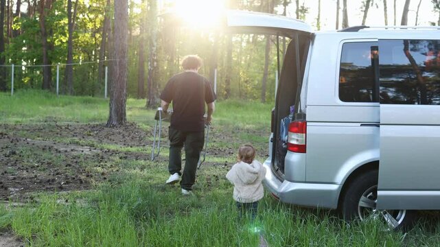 A father sets up a small table and children's toys on the grass in the forest together with his child. Family camping near a camper van