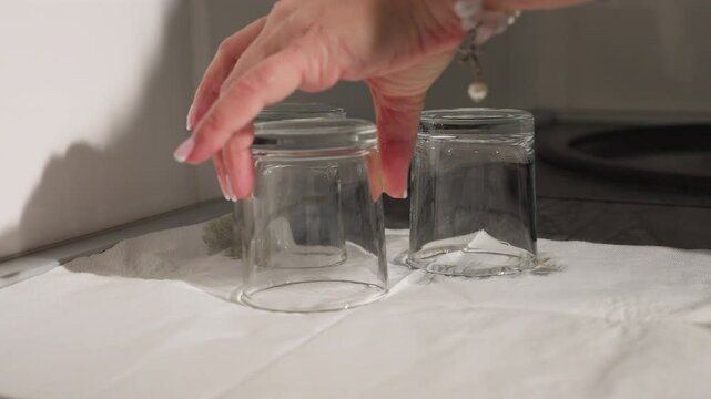 Two clean glass cups placed upside down on white tissue to dry on countertop with third glass cup being carefully positioned under bright kitchen lighting