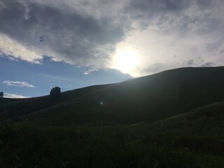 Sunlight peeks through clouds over rolling green hills at dusk near a serene landscape
