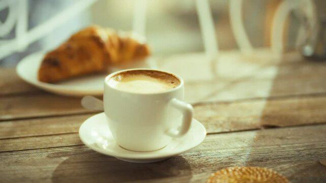 A freshly brewed cup of coffee sits on a small plate beside a flaky croissant in a warm, inviting cafe. Morning sunlight streams in, creating a peaceful atmosphere perfect for enjoying breakfast