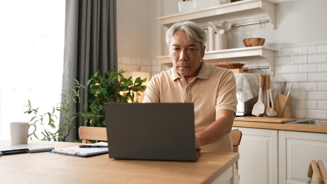 Senior man with casual cloth working on laptop while sitting at home. Elder businessman writing report and looking at financial data while making decision for investment in start up project. Myrmidon.