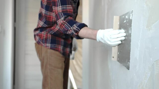Unknown male construction worker wearing protective gloves carefully spreading plaster across wall surface, smoothing uneven areas during home renovation work