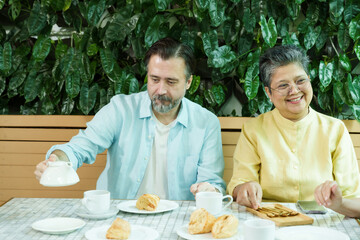 Asian senior woman sits smiling at a cafe table with a Caucasian man pouring tea, both enjoying pastries and creating a joyful, warm atmosphere of friendship and relaxed retired leisure together.