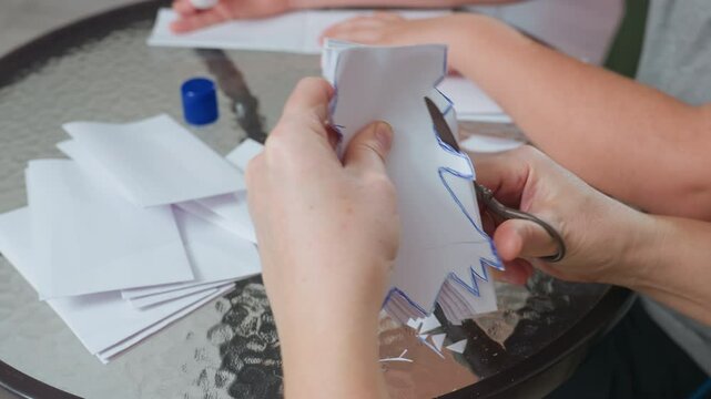 hands engaged in creative process cutting white paper with scissor forming craft design while another hand applies glue on paper on glass table surrounded by scattered paper pieces