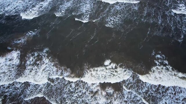 Top down drone view of Baltic Sea waves breaking into white foam over dark coastal water, with rolling surf and flowing patterns creating a natural seascape background moving water and surface texture