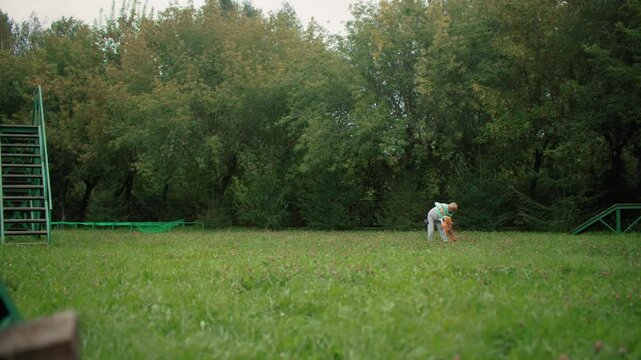 Elegant woman in sportswear playing with dog on grassy field, rolling yo yo toy for excited pet to jump and catch while enjoying active outdoor time surrounded by lush green trees and open space