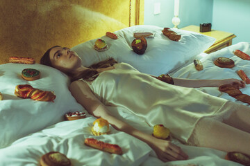 Young woman in silk nightdress lying in bed surrounded by pastries. Surreal and dreamy concept of indulgence, loneliness, and absurdity in a vintage bedroom setting.