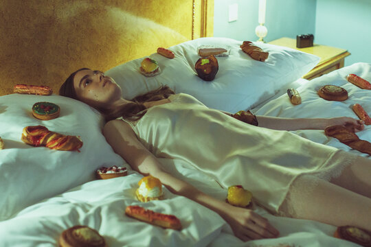 Young woman in silk nightdress lying in bed surrounded by pastries. Surreal and dreamy concept of indulgence, loneliness, and absurdity in a vintage bedroom setting.