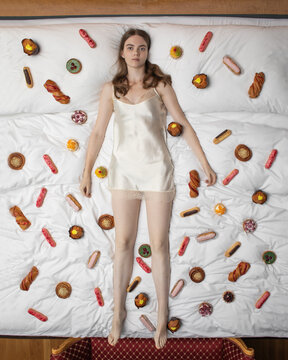 Young woman in silk slip dress lying on a bed covered with assorted pastries. Surreal concept, flat lay composition, mix of fashion, food, and dreamlike aesthetics.