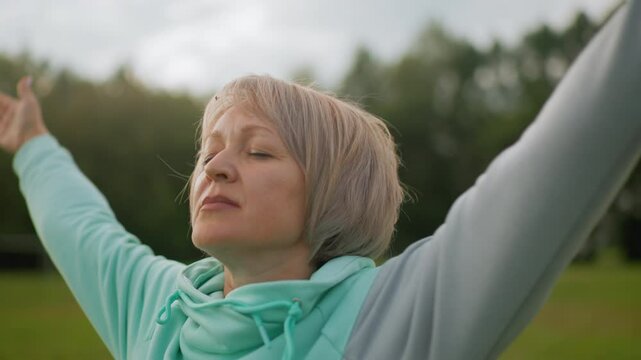 Fair skinned lady with short blond hair wearing blue hoodie practicing controlled breath exercise outdoors eyes closed arms stretched in peaceful green park under soft daylight enjoying tranquility