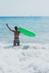 Cheerful girl in floral swimsuit with green inflatable ring raising arms while standing in foamy sea waves on sunny beach, enjoying summer freedom and adventure concept travel, vertical