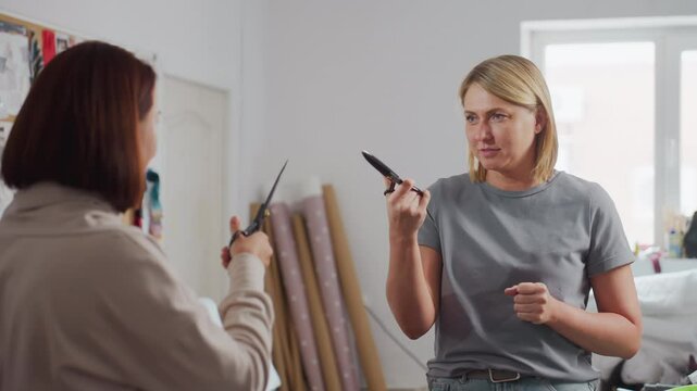 Clothing designers laughing and pretending to sword fight with large sewing scissors in lively bright creative workspace surrounded by colorful textile rolls, design boards, and sewing materials