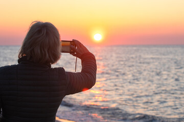 Close-up of a woman capturing a sunset with her smartphone—highlighting connection with nature,...