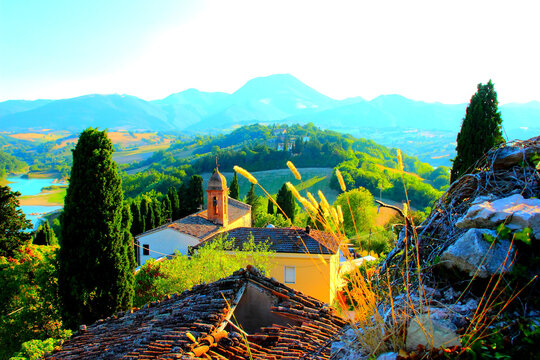 Gorgeous scenery from Lago di Cingoli, rocks and vegetation, an old-fashioned damaged roof, a bell tower, trees, buildings, hills, and the impressive blue Sibillini Mountains in the distant background