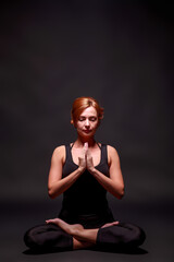 Young slim athletic girl with red hair in black mauka and black shorts doing yoga on black studio background