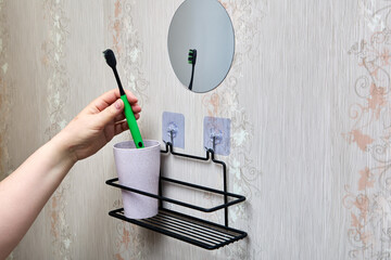 Hand puts green toothbrush into cup on black wire bathroom shelf mounted with transparent nano adhesive hooks for organized bathroom storage.