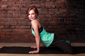 Young slender athletic girl in a turquoise tank top and dark leggings doing yoga against the background of a brick wall
