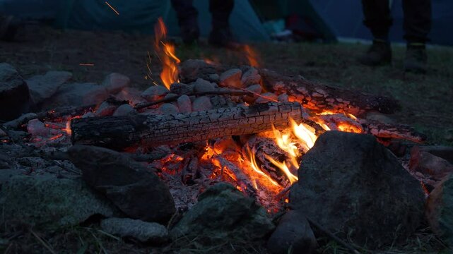 CLOSE UP: Crackling campfire in early evening with glowing embers and charred logs surrounded by stones. Two unrecognizable people stand by the fireplace positioned in the middle of a camping ground.