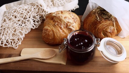 Croissant, netted bag, wooden spoon rest on rustic wooden board beside dark jam jar. Scene emphasizes artisanal breakfast with fresh, simple ingredients for cozy, relaxed morning.